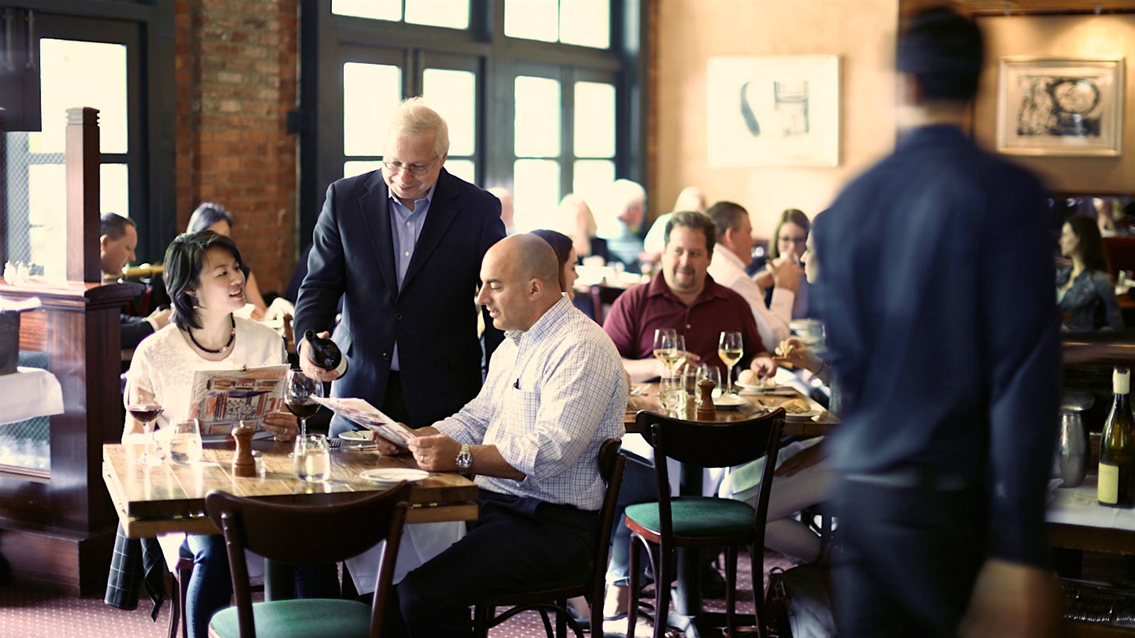 People dining in the Tribeca Grill dining room, with wine director David Gordon pouring a bottle of wine for two diners